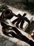 A roll of dark brown velvet ribbon tied around a white gift box with a gold floral pattern, set on a marble surface with pine branches and holiday decor.