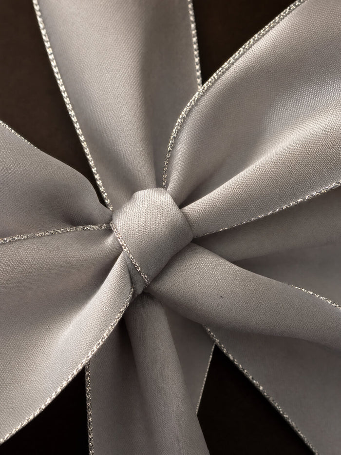 A close-up of a light gray satin ribbon tied into a bow, showing its smooth texture and delicate silver edge trim against a dark background.