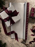 A styled flat-lay of wrapping paper products on a marble-like surface, including a silver gift box tied with a burgundy ribbon, a roll of silver wrapping paper, a roll of burgundy patterned wrapping paper, scissors, ribbon scraps, and another wrapped gift.