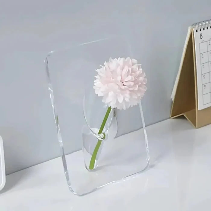 A minimalist Acrylic Frame Vase in clear acrylic holding a single delicate pale pink flower, displayed on a white surface near a desk calendar for home interior decor.