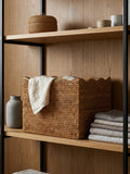 Natural rattan scalloped storage basket styled on wooden shelving with folded linens and ceramics, decorative shelf storage solution