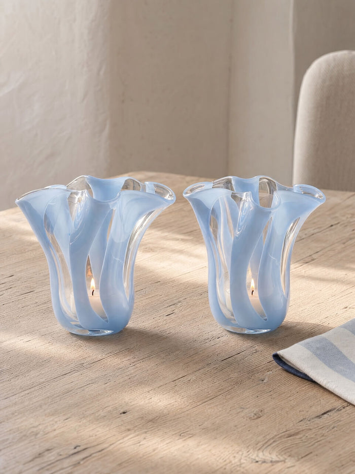Two light blue fluted glass candle holders with a wavy, ribbed design, shown on a light wood table in a softly lit interior setting.