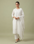 A full-body studio portrait of a South Asian female model with her hair tied back in a neat bun, wearing a timeless white cotton Chikankari kurta suit set. The ensemble includes a long, straight-cut kurta with subtle tonal embroidery, matching white straight pants, and a soft white dupatta with delicate lace trim. She is posed in a graceful three-quarter profile against a solid, soft sage-green background, looking gently towards the camera with a soft smile. Her right hand is raised delicately near her ches