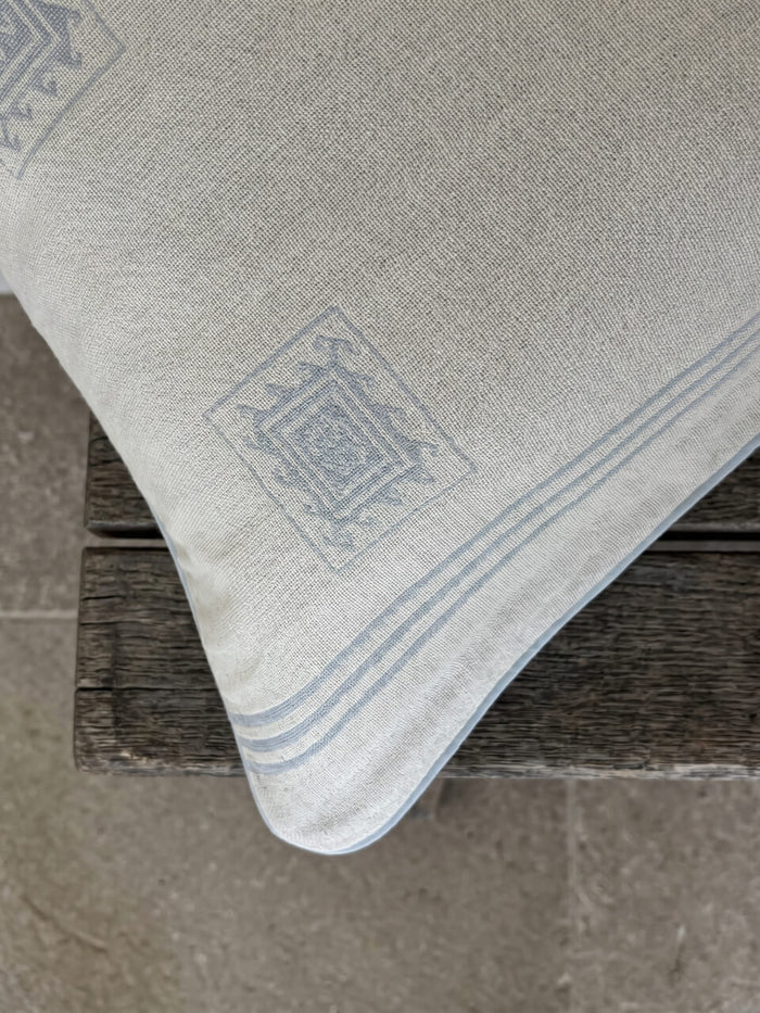 Close-up detail of a luxury cream and blue patterned BCI cotton cushion cover with woven geometric detailing and soft blue piping, styled on rustic timber for a relaxed contemporary aesthetic.