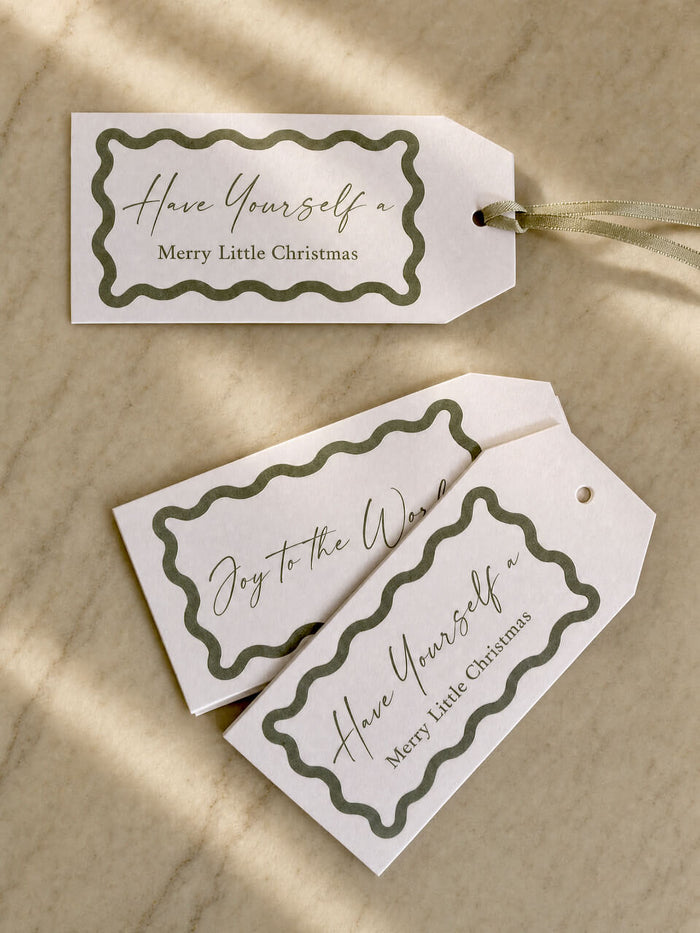 Three decorative gift tags with scalloped olive green borders and cursive holiday text, arranged on a light beige marble surface with soft shadows.