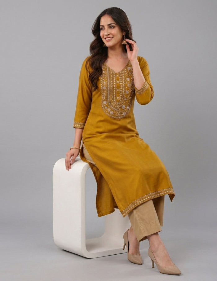 A woman models a mustard yellow kurta with gold embroidery on the chest and hem, paired with beige trousers and beige heels, sitting on a white modern stool against a gray background.