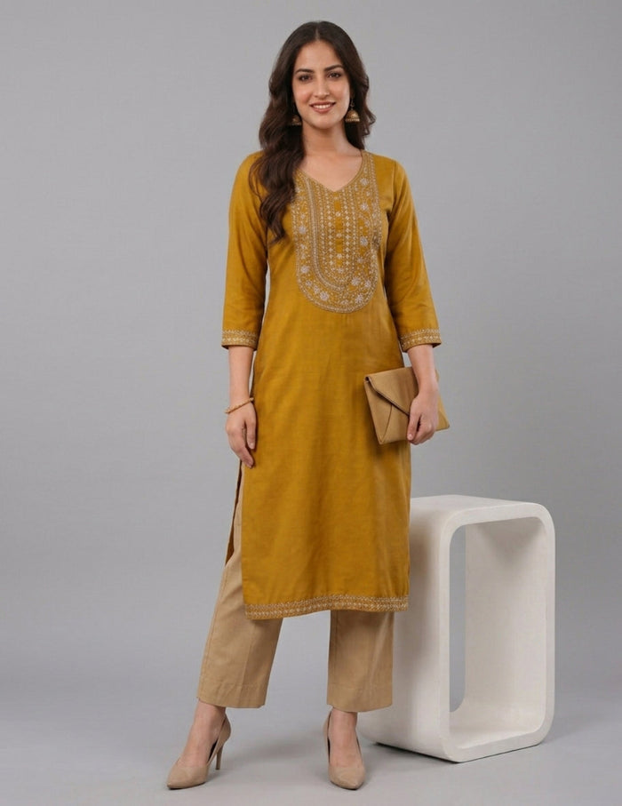 A woman models a mustard yellow kurta with beige embroidery on the chest and sleeve cuffs, paired with beige trousers, standing next to a white modern block stool against a gray studio background.