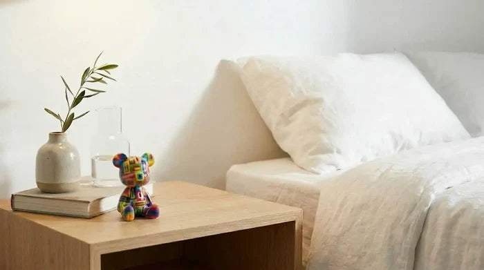 Colorful handcrafted bear figurine on wooden nightstand beside white bedding and vase with greenery
