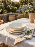 Cream scalloped dinnerware set including dinner plate, side plate, and pasta bowl styled on a white linen table outdoors with olive branches and Mediterranean garden surroundings.