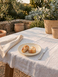 Cream scalloped side plate styled with rustic bread on a white linen tablecloth outdoors, surrounded by terracotta pots and Mediterranean garden greenery in warm evening light.