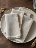 Four folded light putty linen napkins with frayed edges, arranged on a round ceramic plate on a dark wooden table, with a gold fork and glass visible.
