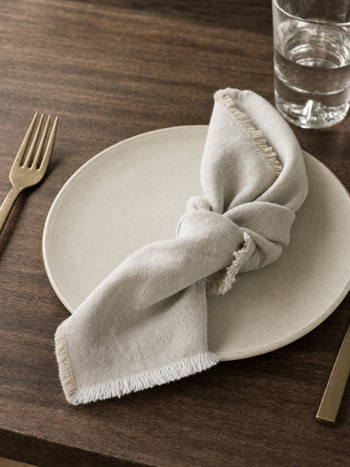A light putty linen napkin with frayed edges, tied in a knot on a round ceramic plate, set on a dark wood table with a gold fork and a glass of water.