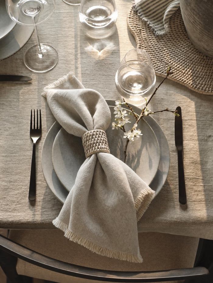 A styled table setting featuring a light Putty linen napkin tied with a woven white rattan napkin ring, layered over Luso plates on a putty linen tablecloth, with silver cutlery and glassware.