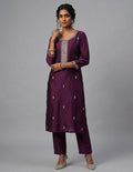 A woman modeling a deep purple Indian ethnic outfit consisting of a long kurta with silver embroidery and matching trousers, paired with silver high heels, against a solid gray studio background.