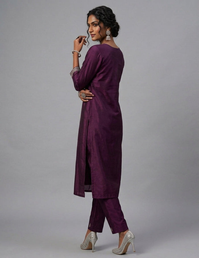 A woman modeling a deep purple two-piece kurta and trouser set with silver embellished high heels, photographed against a solid gray studio background.